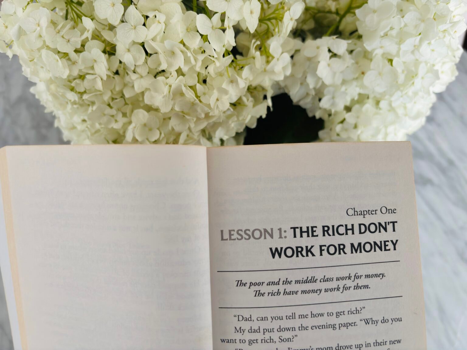 Open book Rich Dad Poor Dad beside white flowers – Chapter 1: The Rich Don’t Work for Money