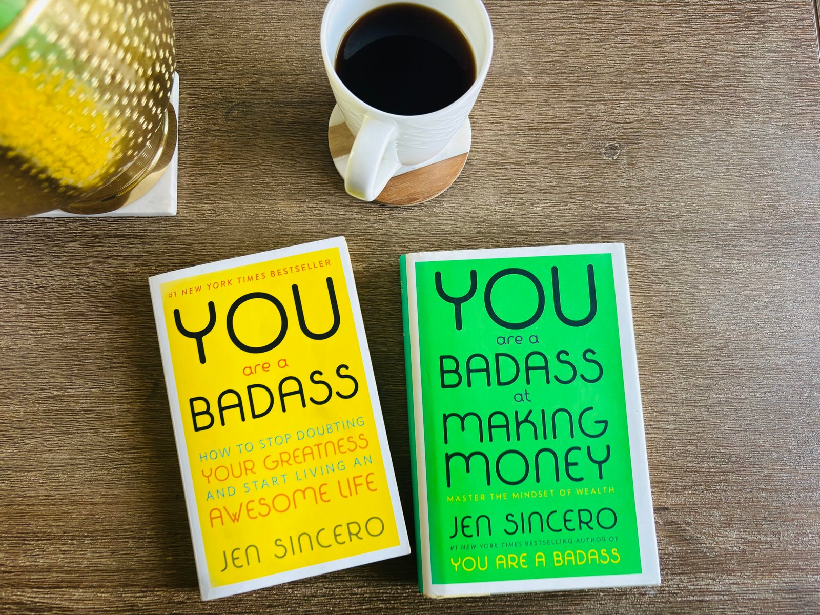 Two Jen Sincero books — You Are a Badass and You Are a Badass at Making Money — on a cozy wooden table with a cup of black coffee