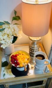 Warm bedside lamp beside coffee, fruit bowl, and flowers on a mirrored table