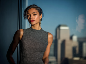 A confident woman in a sleek dress standing by a window with city views, symbolizing self-belief and inner conviction in creative entrepreneurship.